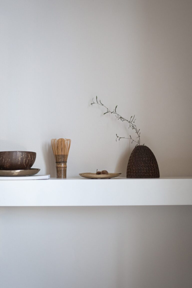 A minimalist white shelf displays a wooden bowl, a bamboo whisk, a small plate, and a textured vase with a delicate twig against a plain light-colored wall.