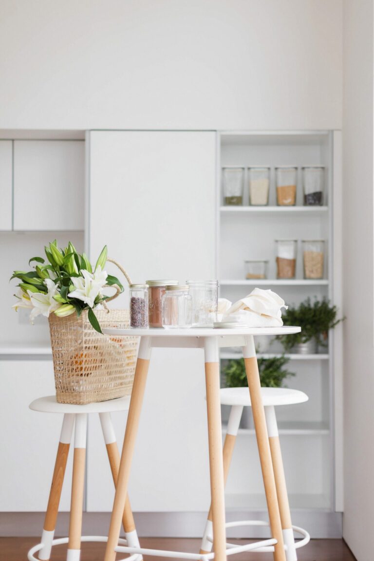 A modern kitchen with wooden stools, a round white table holding glass jars, and a wicker basket of white lilies. Shelves in the background display jars filled with grains and herbs.