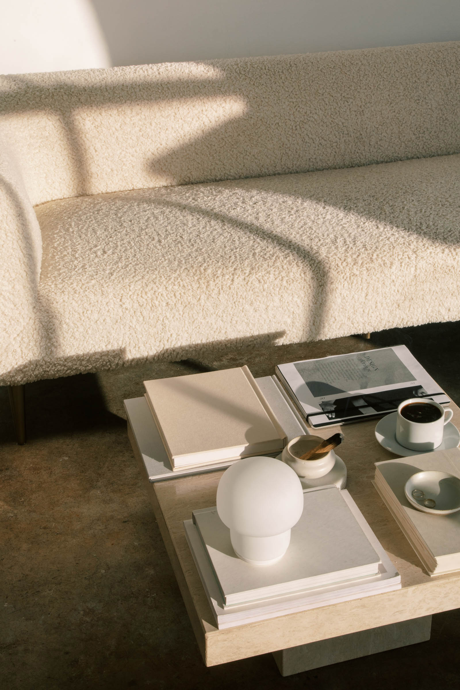 A modern beige sofa sits beside a low coffee table with books, a small white lamp, a newspaper, and two cups of coffee, bathed in warm sunlight and soft shadows.