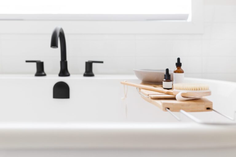 A white bathtub with black fixtures and a bath tray holding bottles, a brush, a bowl, and a sponge, set in a bright, minimalistic bathroom.