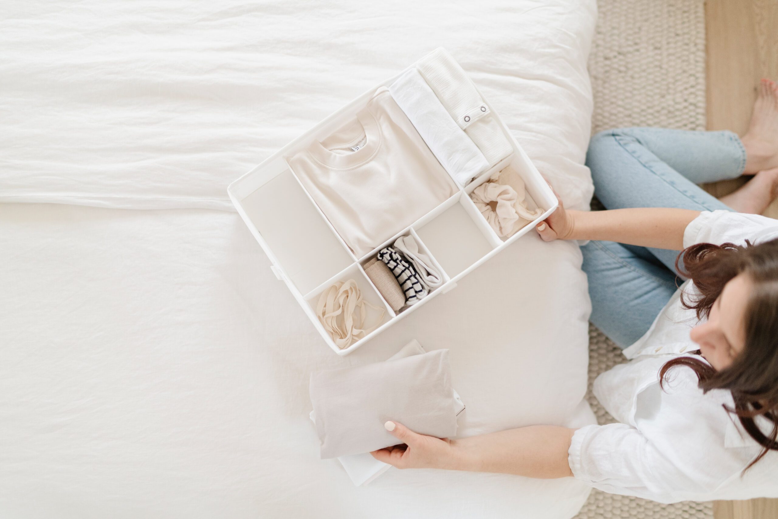 A person sits on the floor beside a bed, organizing neatly folded clothes in a white drawer organizer filled with light-colored garments.