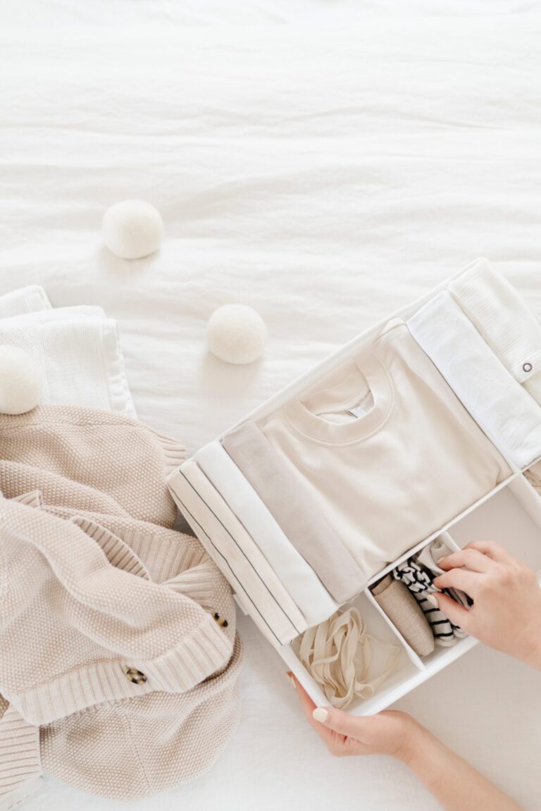 A hand organizes neatly folded neutral-colored clothes in a white drawer, with folded sweaters and wool dryer balls on a white bedspread nearby.