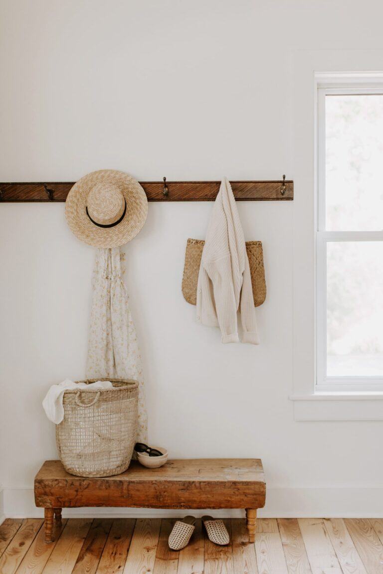 A wooden bench sits below a wall rack with hooks holding a straw hat, patterned scarf, cream sweater, and a woven bag. A basket with a blanket and a pair of slippers are on the bench. A window lets in natural light.