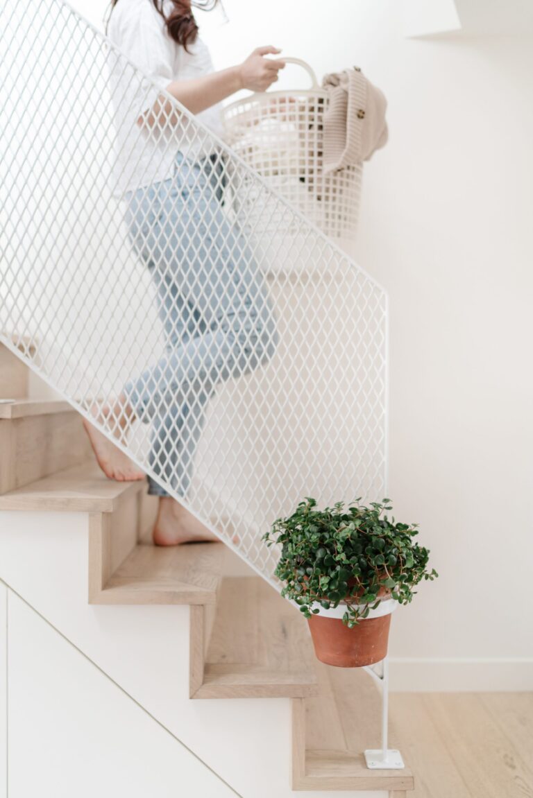 A barefoot person in jeans carries a laundry basket down wooden stairs with a white mesh railing. A potted plant is attached to the side of the railing.