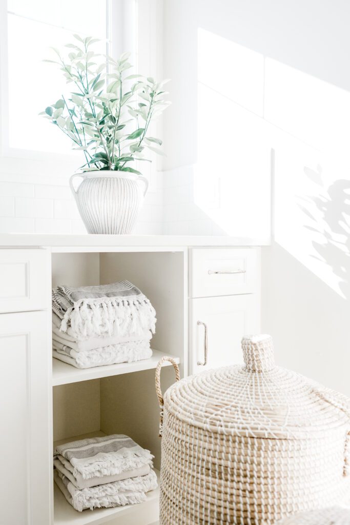 Bright laundry room with white cabinets, a woven basket with lid, neatly folded towels on shelves, and a white vase with green leafy branches near a sunlit window.