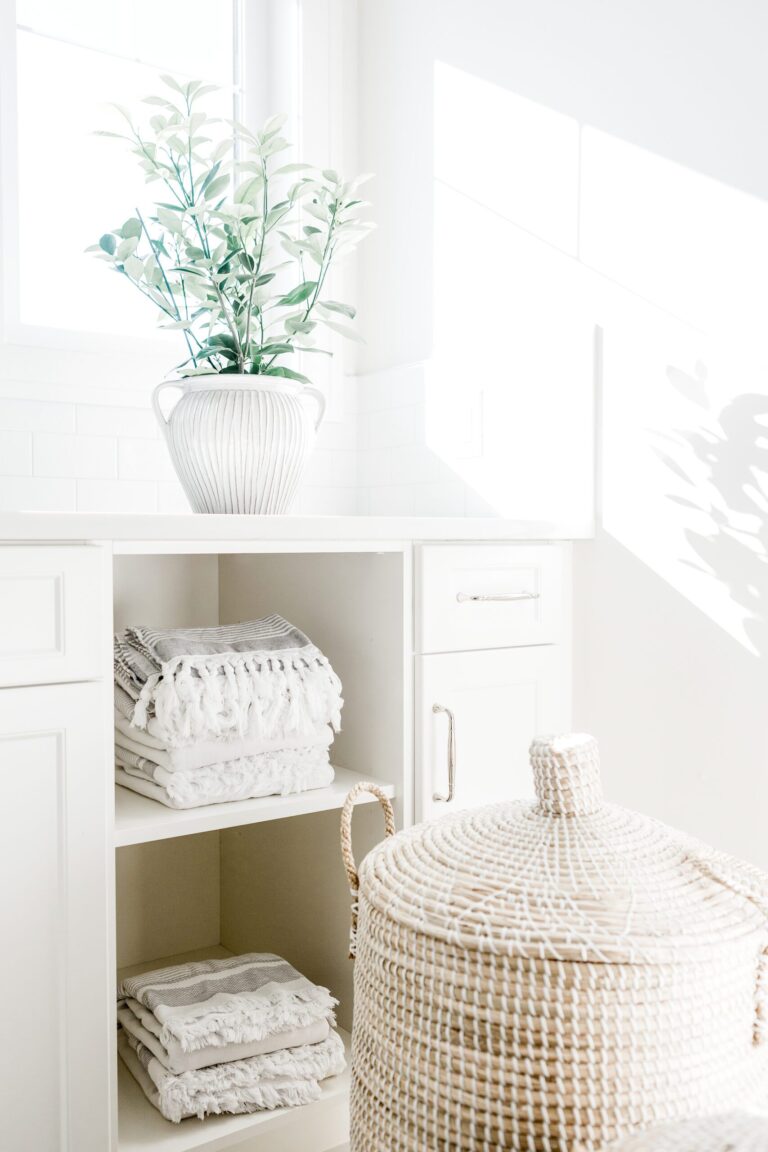 Bright, airy laundry room with white shelves holding neatly folded towels, a woven lidded basket in the foreground, and a plant in a white vase placed on the counter by a sunny window.
