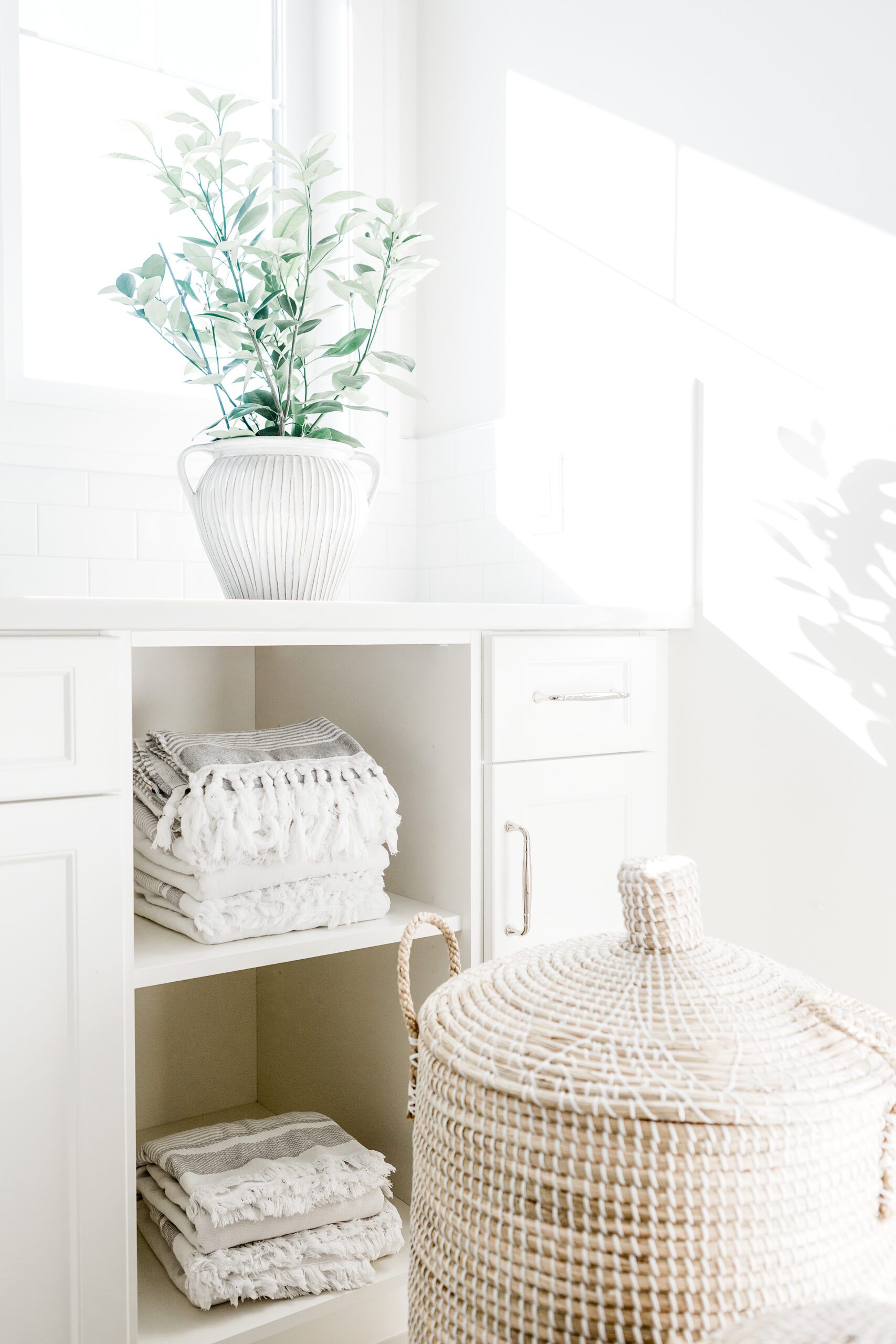Bright, airy laundry room with white shelves holding neatly folded towels, a woven lidded basket in the foreground, and a plant in a white vase placed on the counter by a sunny window.