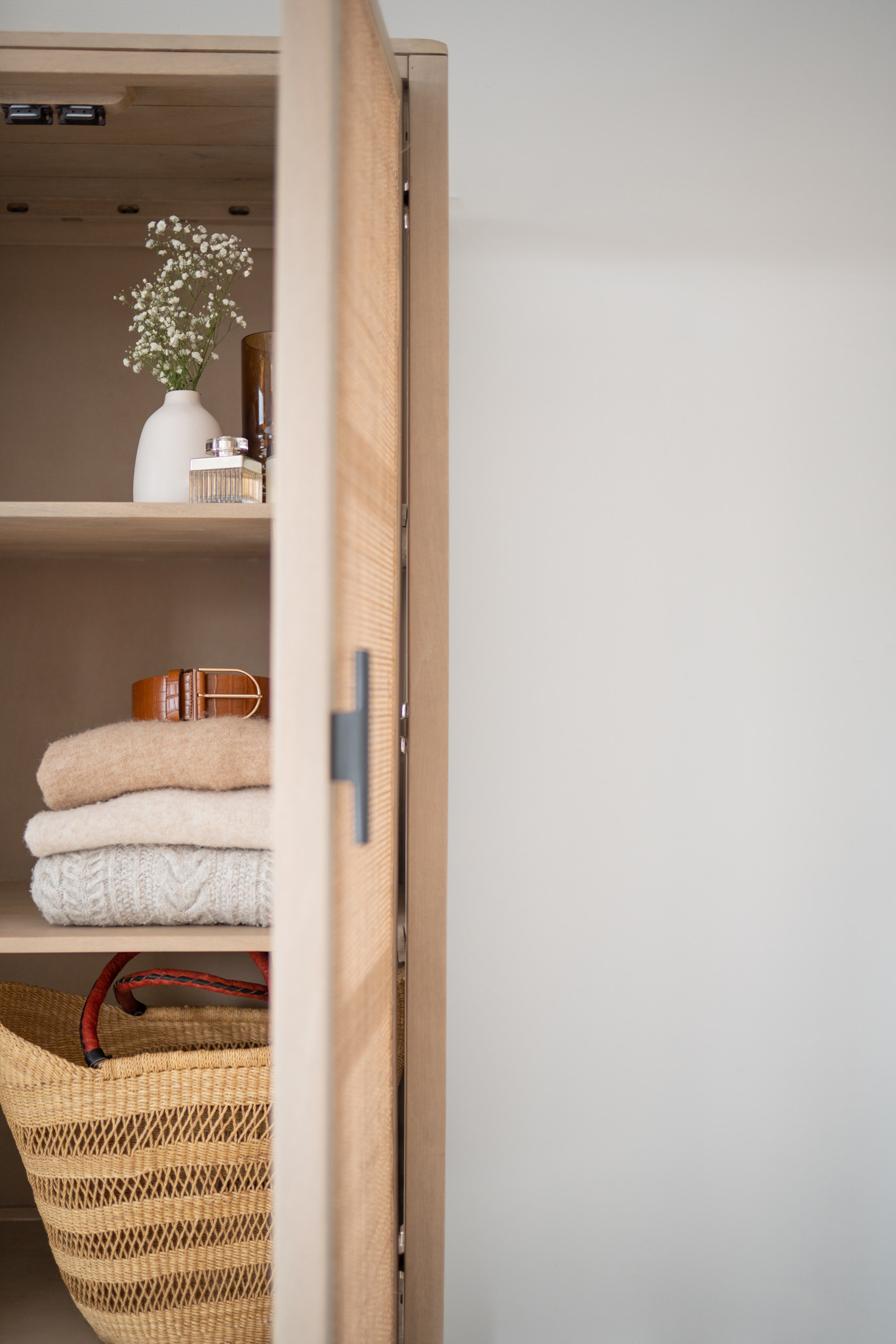 A partially open wooden cabinet reveals folded sweaters, a woven basket, a small vase with flowers, and a few small items on a shelf.