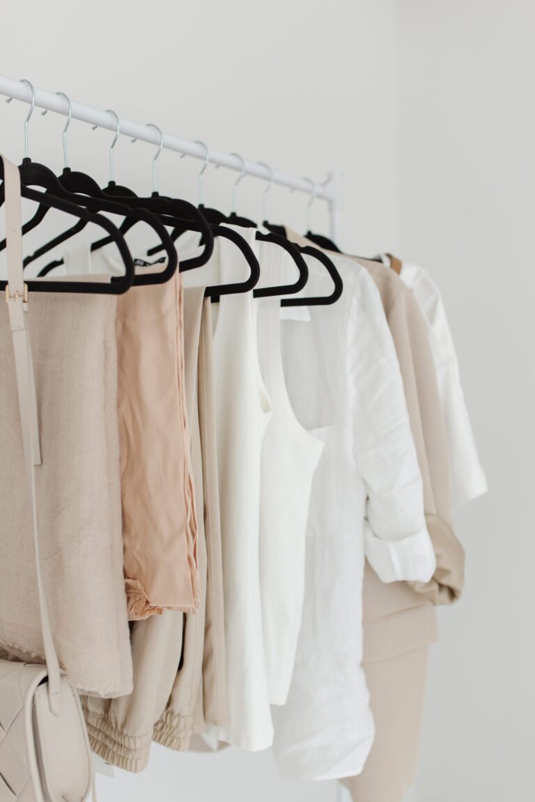 A clothing rack holds beige, white, and tan tops and pants on black hangers, with a beige purse hanging on the side. The background is a bright, minimalist room.