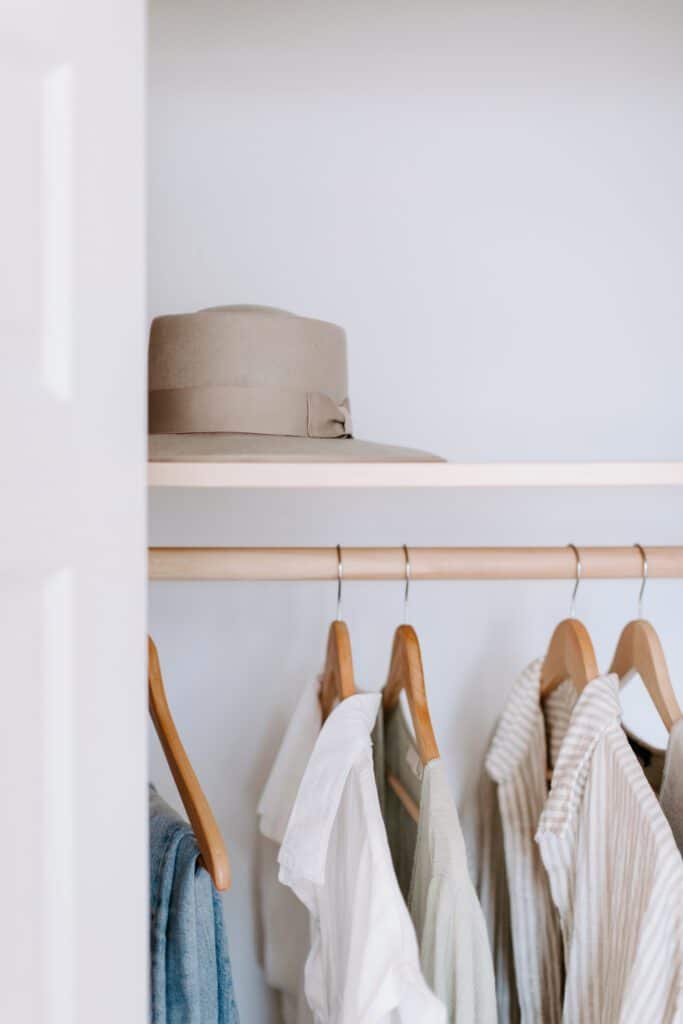A beige hat rests on the top shelf of a closet above a clothes rail with wooden hangers holding light-colored shirts and tops.