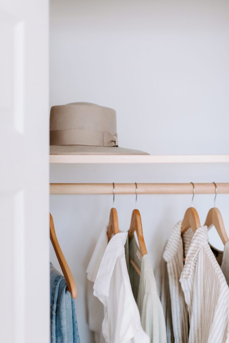 A beige hat sits on a shelf above a row of light-colored shirts hanging on wooden hangers in a minimalist closet.