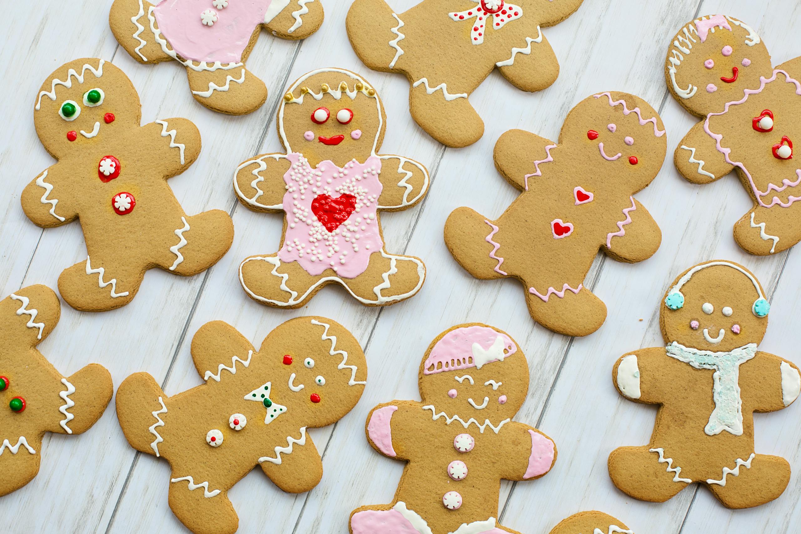 Decorated gingerbread cookies shaped like people are arranged on a white wooden surface, each with different icing designs, buttons, and facial expressions.