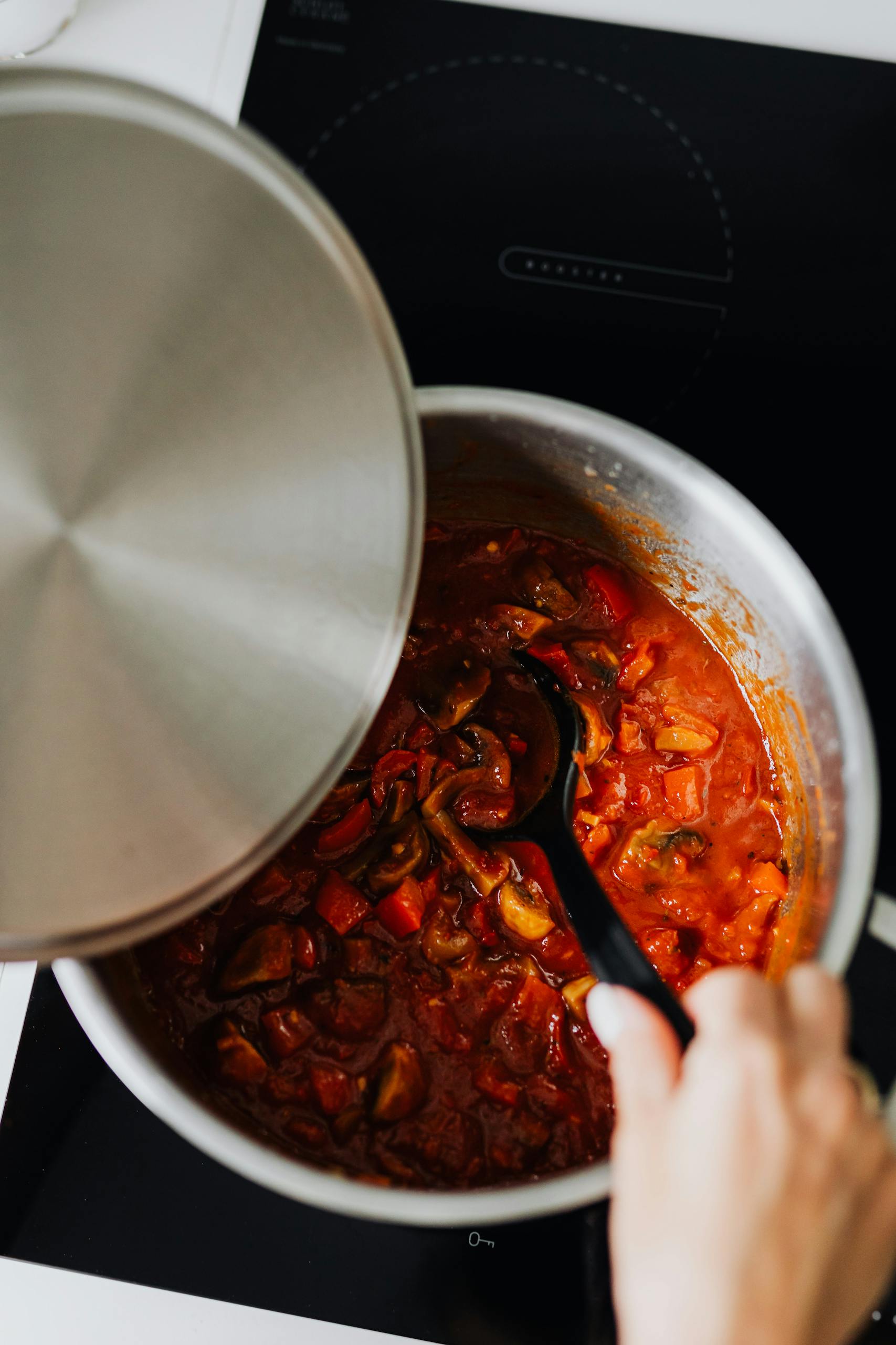 A hand holding a pot lid and a black ladle stirs a tomato-based vegetable stew on a stovetop.