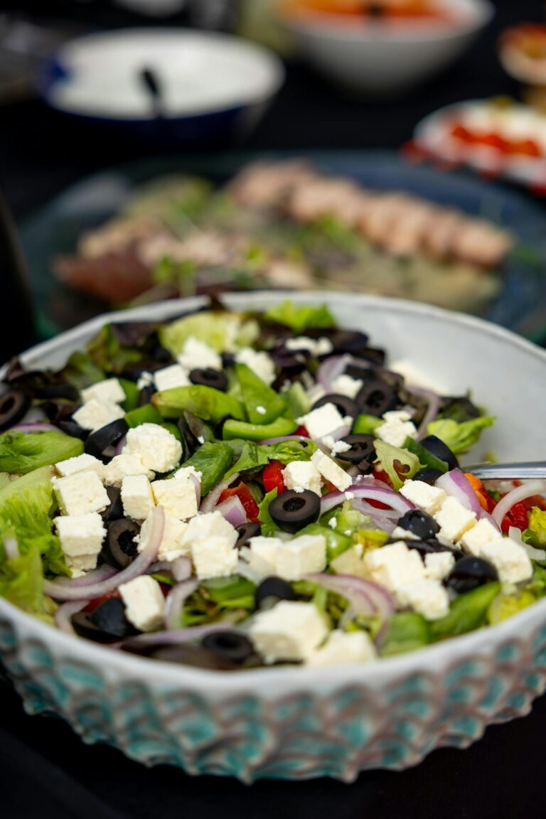 A bowl of fresh salad with lettuce, sliced red onions, black olives, diced feta cheese, and red bell peppers on a table with other blurred dishes in the background.
