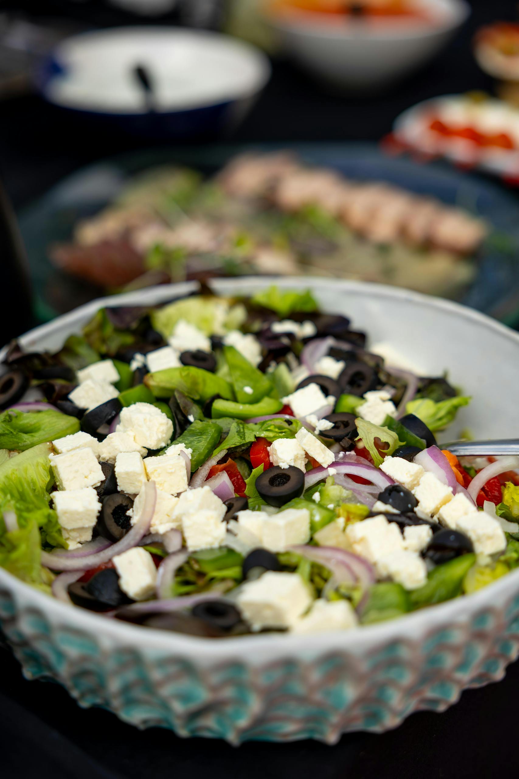A bowl of fresh salad with lettuce, sliced red onions, black olives, diced feta cheese, and red bell peppers on a table with other blurred dishes in the background.
