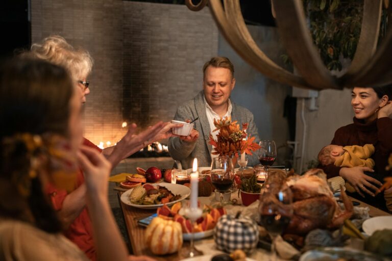 A family gathers around a dinner table filled with food, candles, and autumn decorations. One person passes a dish to another while a woman cradles a baby. A fireplace glows in the background, creating a warm atmosphere.