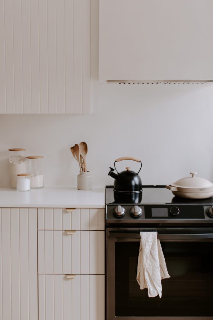 Minimalist kitchen with white cabinets, jars, wooden utensils in a holder, a black kettle, a beige pan on a stove, and a cream-colored towel hanging from the oven handle.