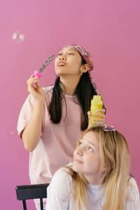 Two young girls enjoy blowing bubbles in a bright studio setup with a pink background.