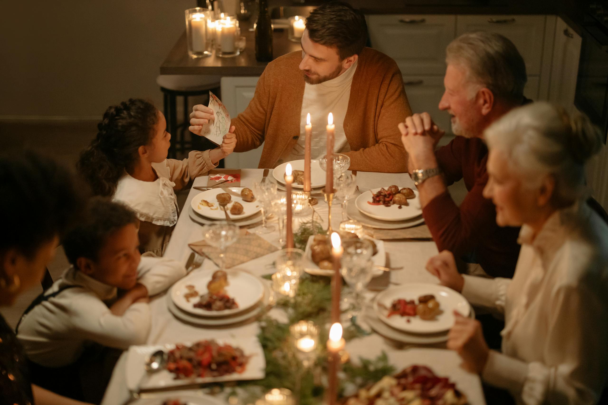 A family sits around a candlelit dinner table, sharing food and smiling. An adult shows something on a phone to a young girl, while others look on warmly. The scene feels festive and cozy.