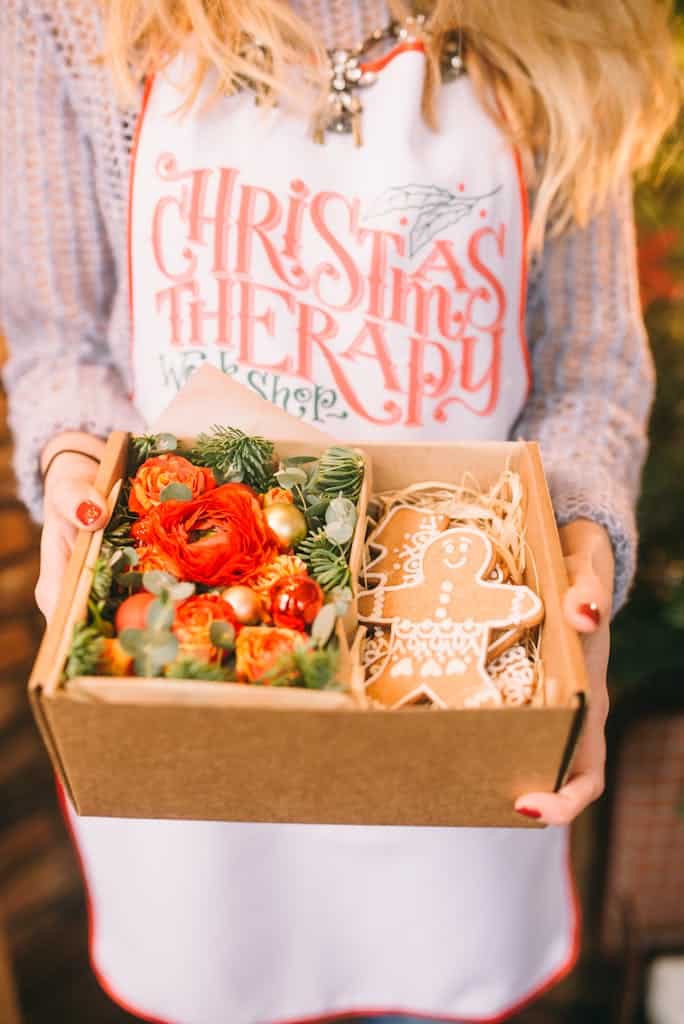 Woman holding a Christmas-themed gift box with gingerbread and flowers, wearing 'Christmas Therapy' apron.
