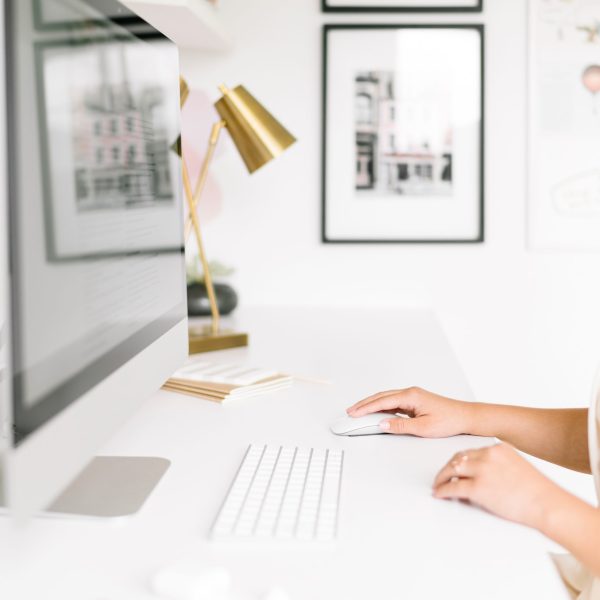 A person using a desktop computer at a white desk with a keyboard and mouse. The workspace is decorated with framed art, a gold lamp, and office supplies, creating a bright, minimalistic atmosphere.